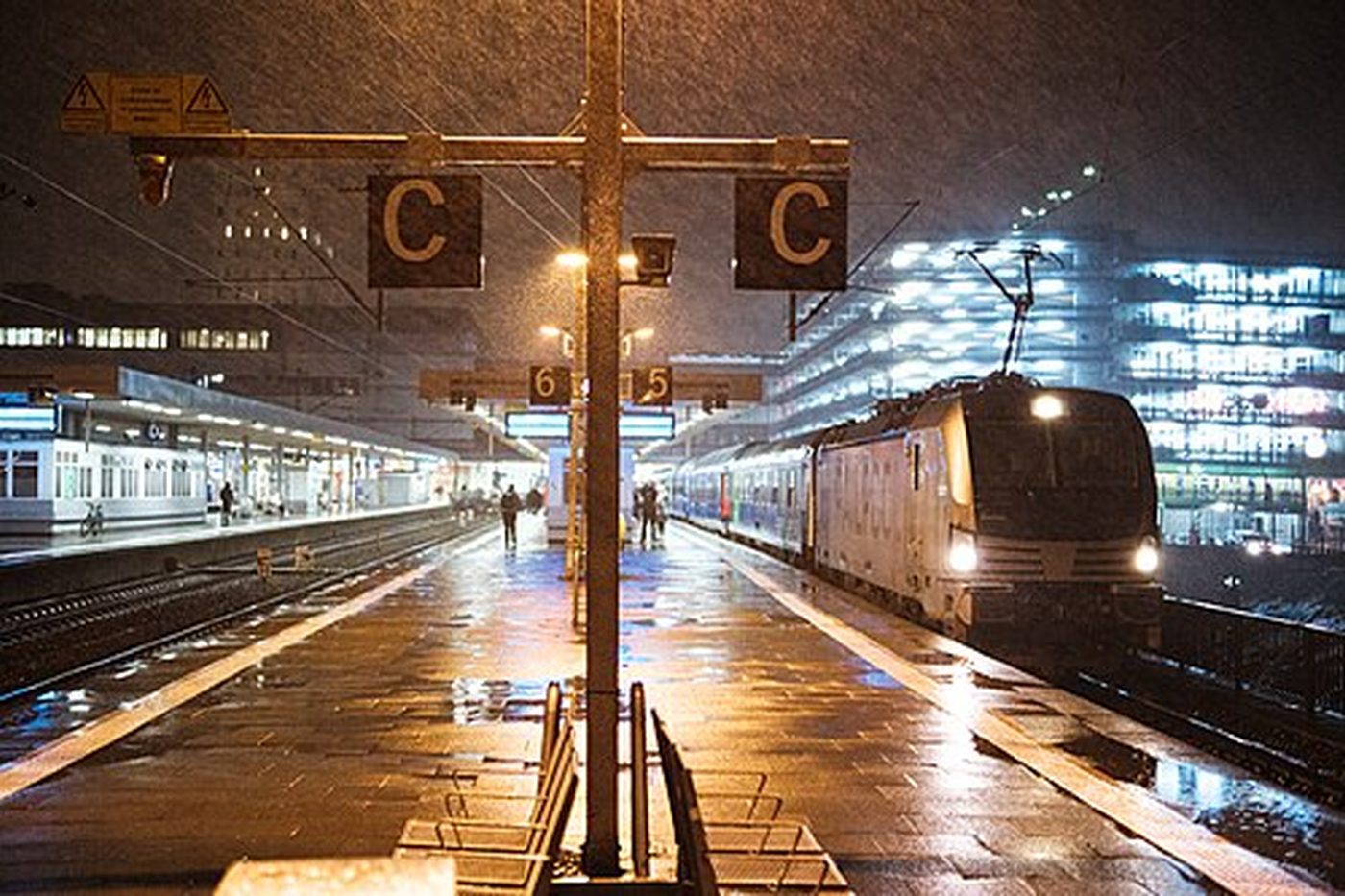 Ein nasser Bahnsteig bei Nacht, Lampen an, ein Zug wartet am hinteren Gleis