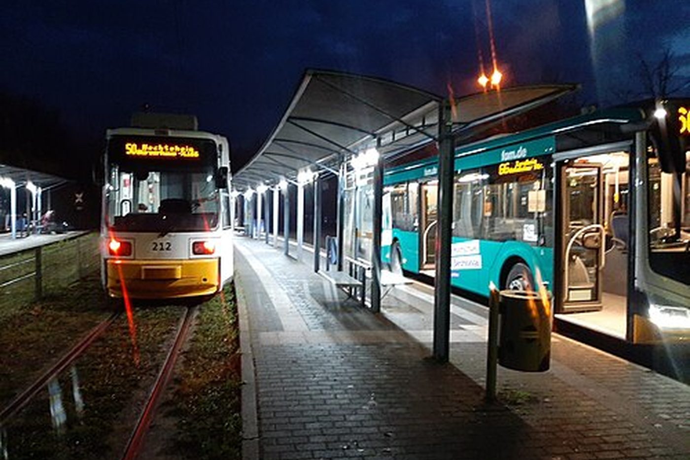Eine Straßenbahnhaltestelle in einer deutschen Stadt bei Nacht mit beleuchtetem Wartehäuschen und wartendem Bus